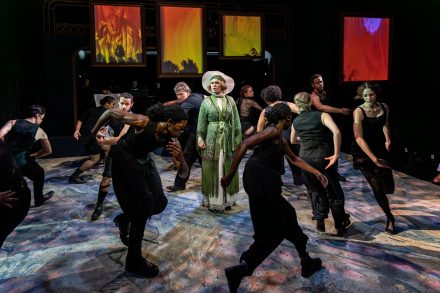 An image of a theatrical performance. A women in a green dress is surrounded by a group of dancers wearing black.