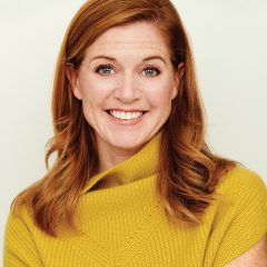 A woman with red hair smiles in a studio headshot, wearing a yellow sweater against a light background.