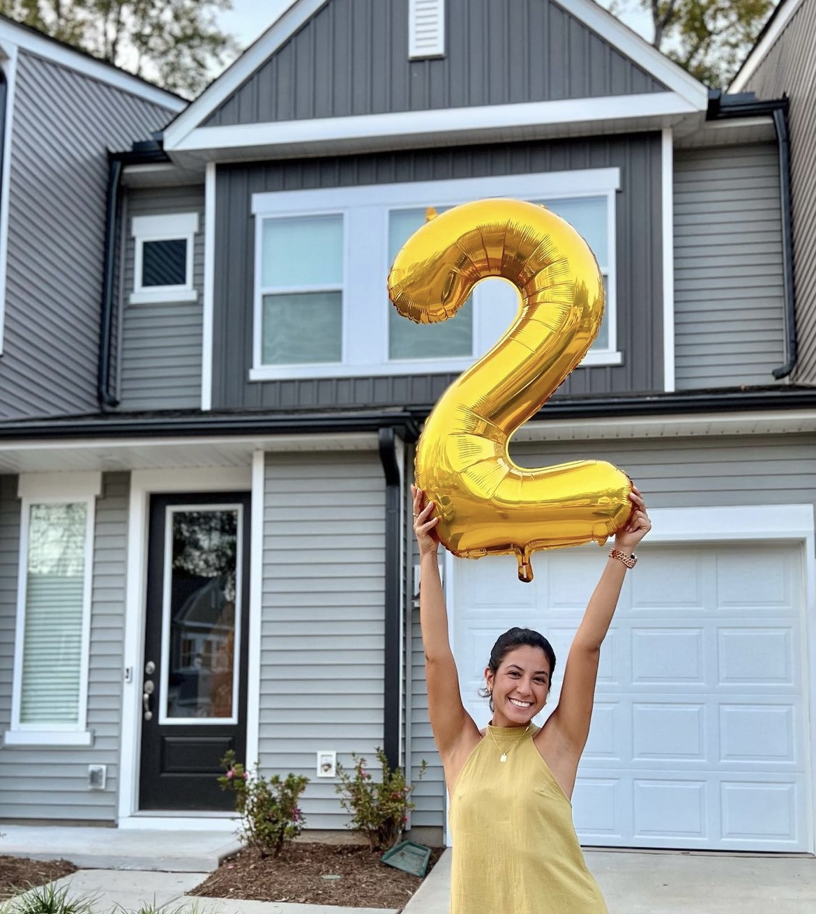 A woman in a yellow shirt holds up a balloon number 2 in front of a house. She is smiling.