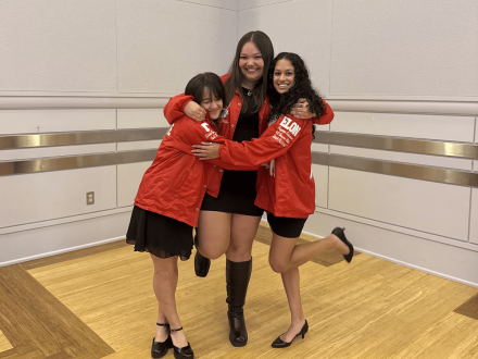 From left to right, Lillian Chen ’27, Reagan Wible ’26 and Anita Bhat ’27 share a joyful moment, embracing one another in coordinated red jackets. The trio smiles and leans in together, capturing a celebratory and spirited pose indoors.