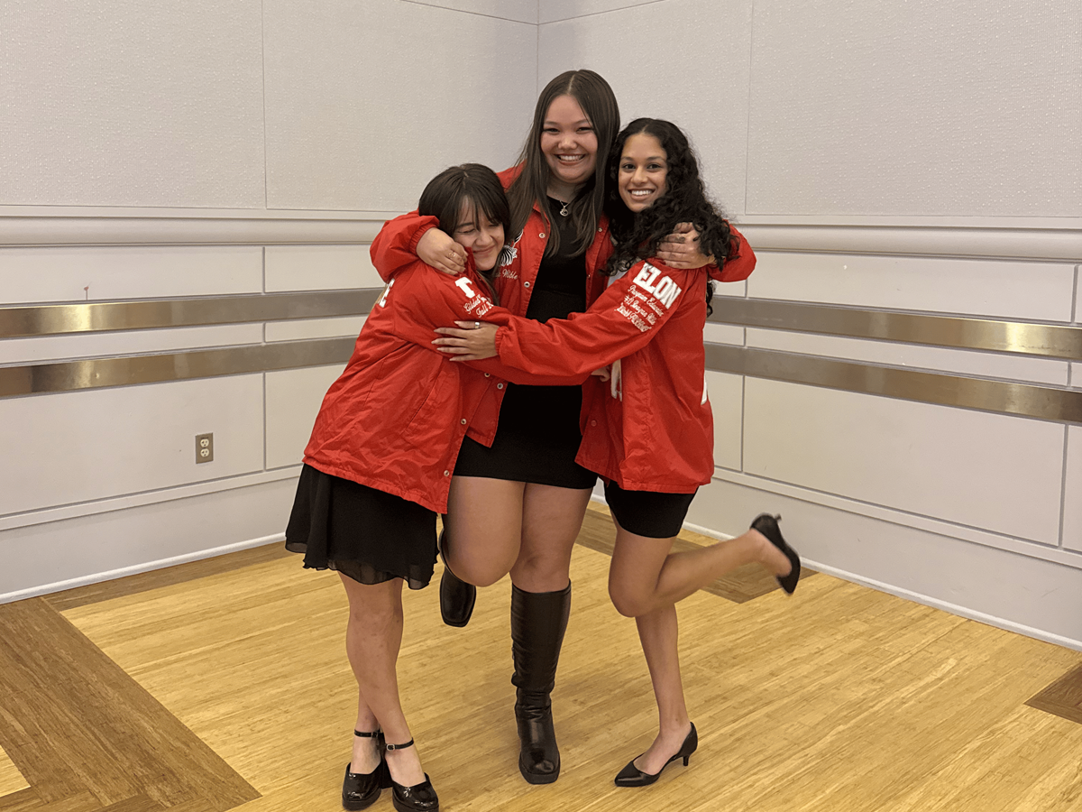 From left to right, Lillian Chen ’27, Reagan Wible ’26 and Anita Bhat ’27 share a joyful moment, embracing one another in coordinated red jackets. The trio smiles and leans in together, capturing a celebratory and spirited pose indoors.