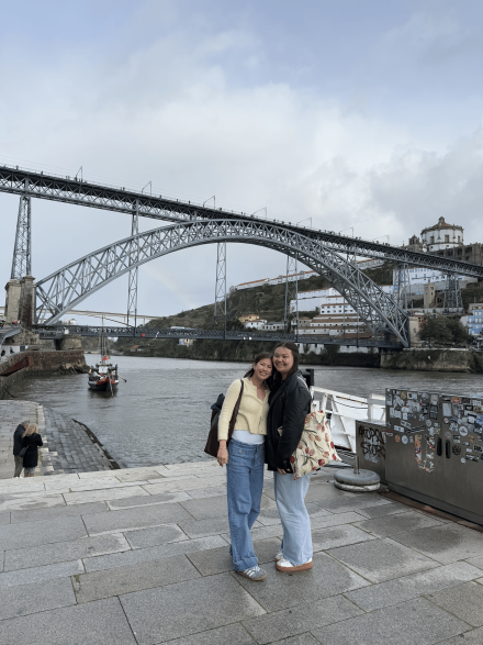 From left to right, Anna Marchesseault ’24 and Reagan Wible ’26 stand together along the waterfront, smiling in front of a sweeping arched bridge. The scenic backdrop and overcast sky frame a relaxed, travel moment as they pose side by side.