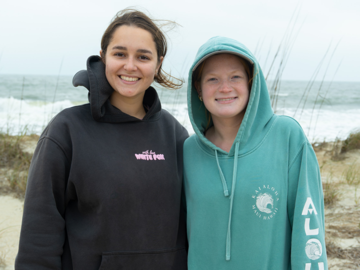 Two students stand smiling together on a beach, wearing hoodies, with ocean waves and sea grass in the background.