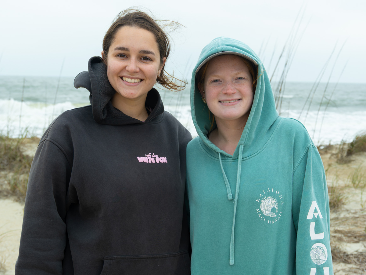 Two students stand smiling together on a beach, wearing hoodies, with ocean waves and sea grass in the background.