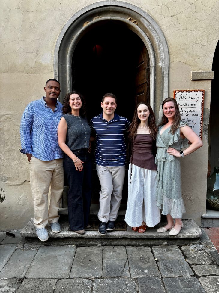 Five people pose for a photo in front of a tan wall with a black door