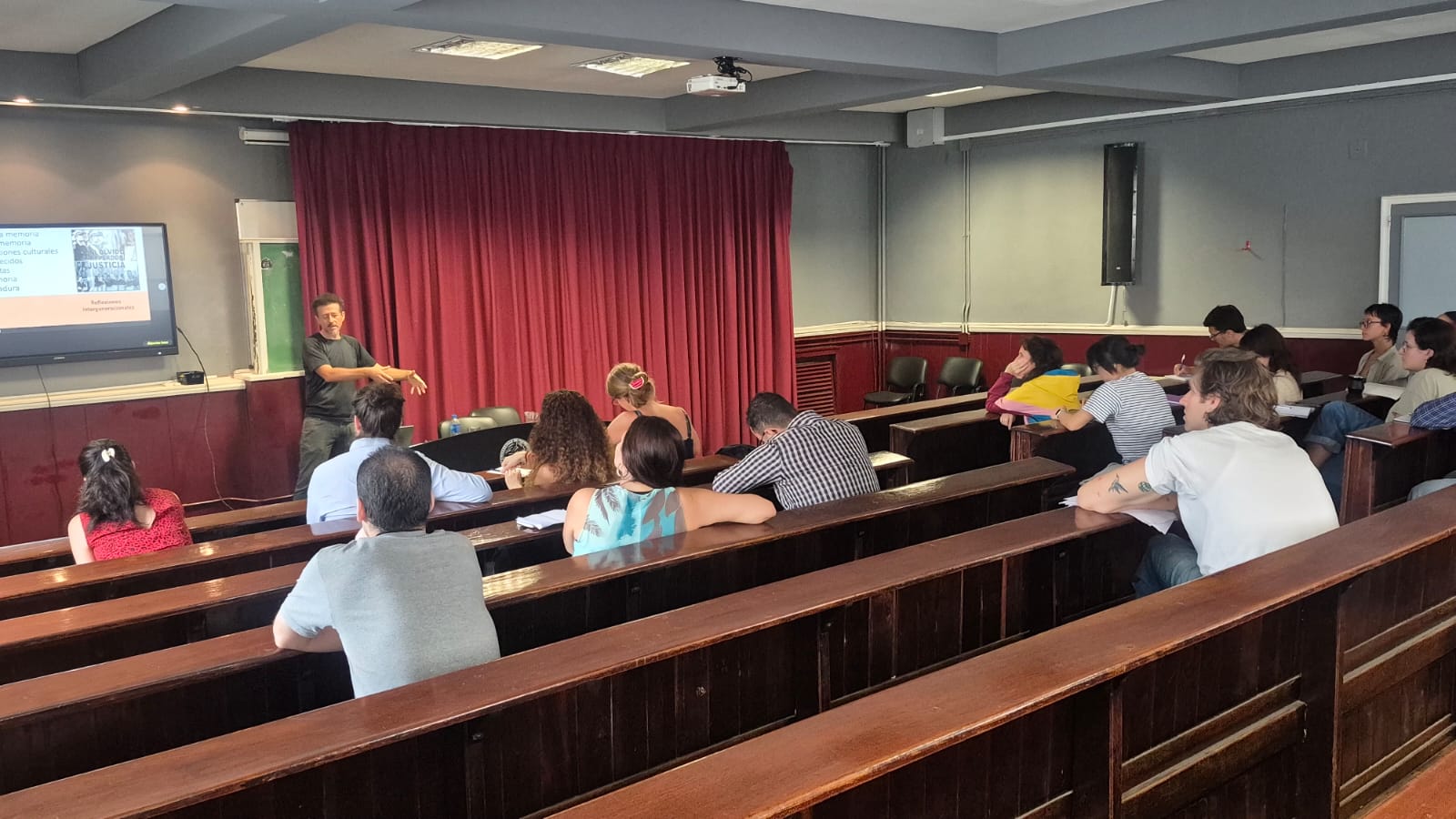A man gives a ledcture at the front of a college classroom as students listen in wooden seats