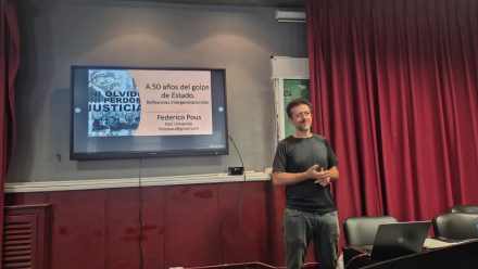 A man stands in front of a TV and red curtain giving a presentation