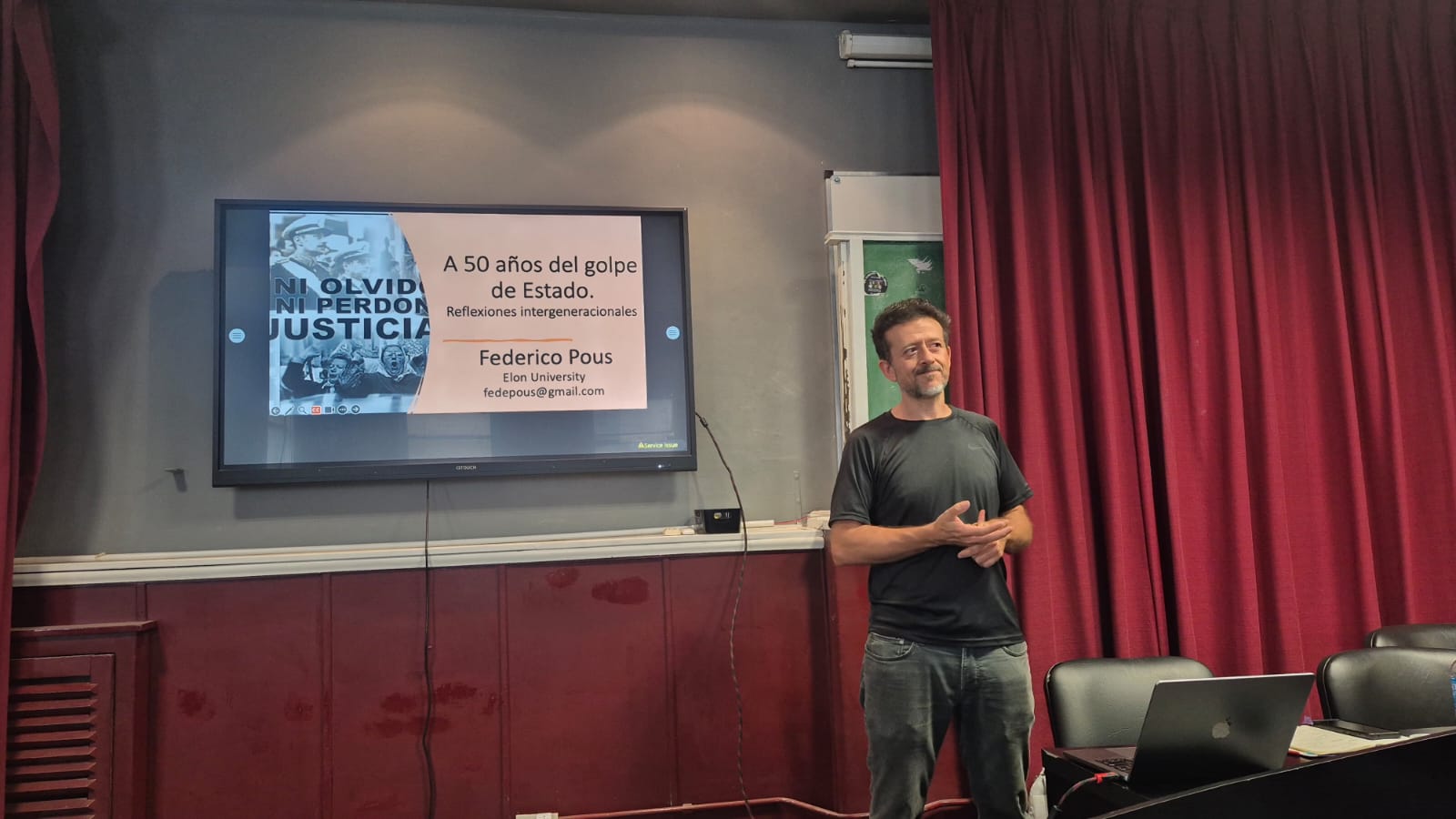 A man stands in front of a TV and red curtain giving a presentation