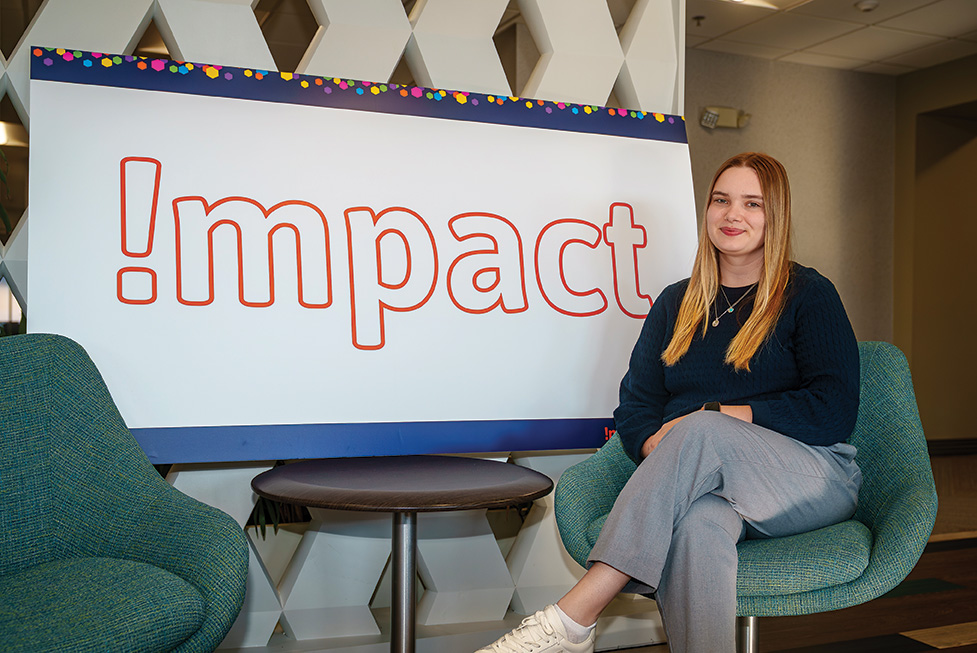A woman sits in a chair beside a large sign reading “Impact” in a modern indoor space.
