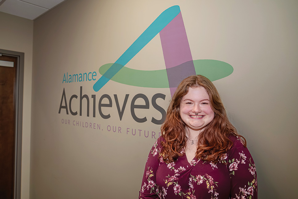 A woman smiles while standing in front of a wall displaying the “Alamance Achieves: Our Children, Our Future” logo.