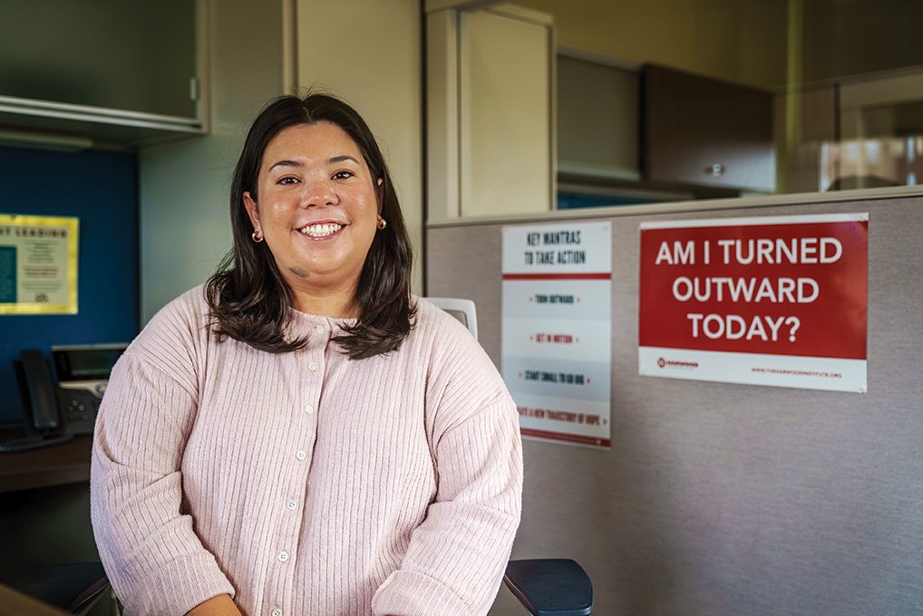 A woman smiles while seated in an office cubicle, with a sign reading “Am I turned outward today?” posted behind her.