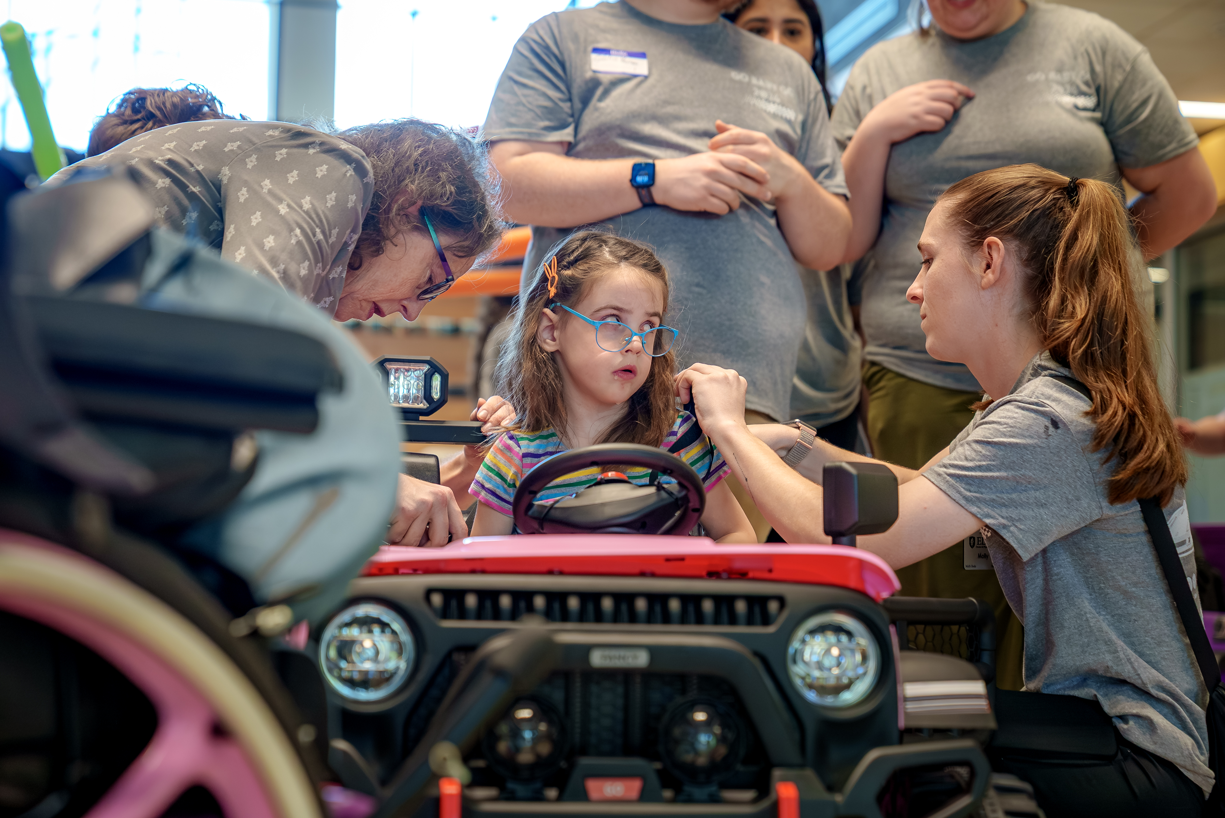Volunteers assist a young girl wearing glasses as she sits in a red adaptive toy car.