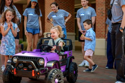 Little girl drives a purple ride-on toy car as other children and adults observe and smile.