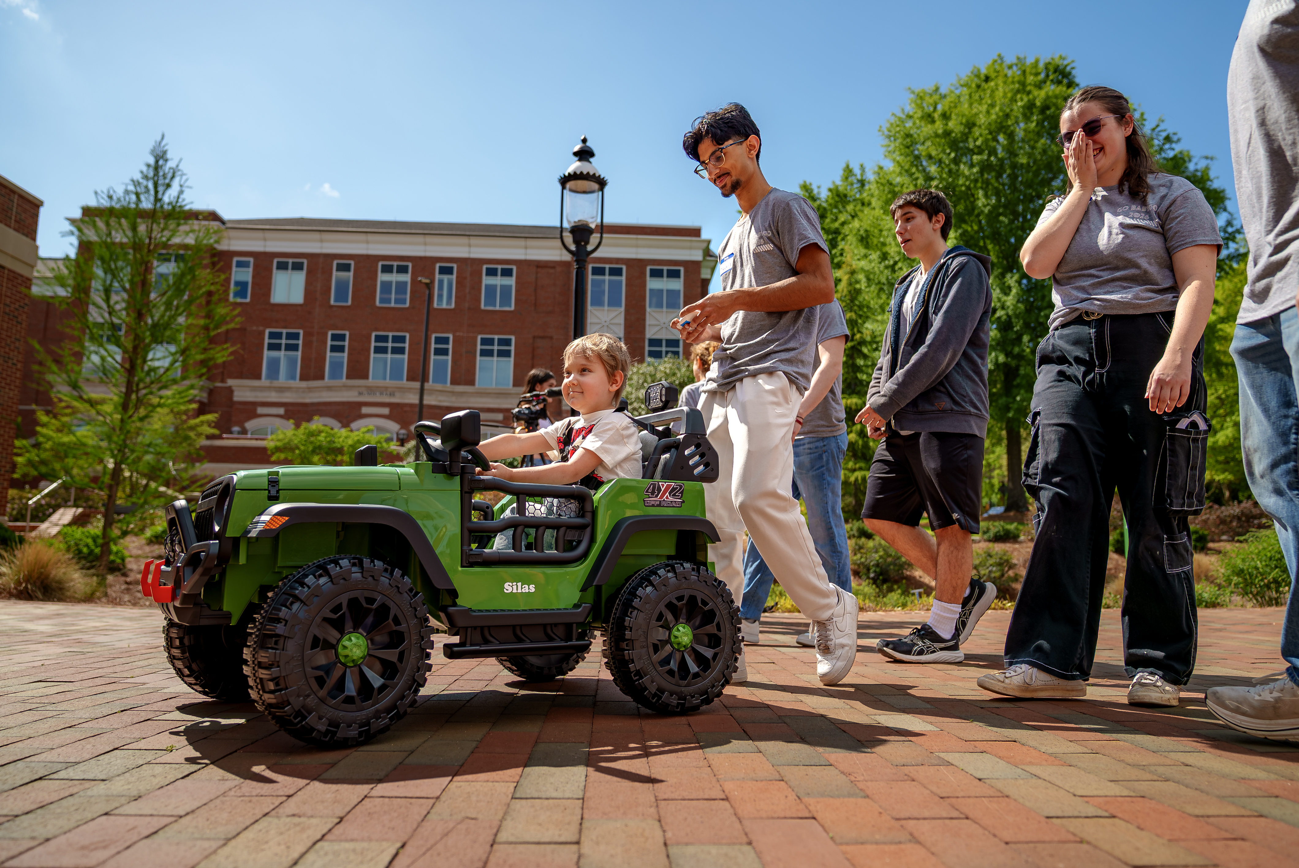  Young child drives a green ride-on toy Jeep on a brick walkway while a group of students walks alongside, smiling and supervising on a sunny campus.