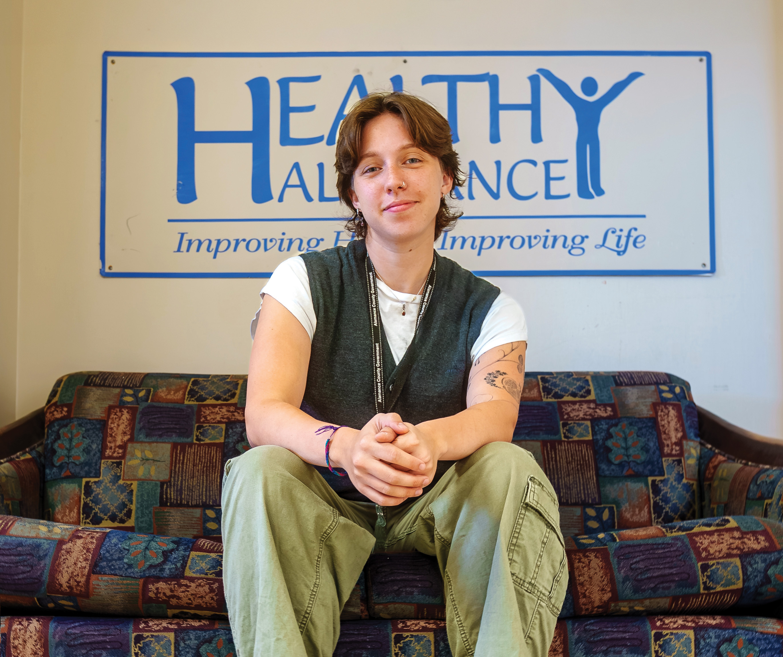 A person sits on a couch in front of a sign reading “Healthy Alamance: Improving Health, Improving Life.”