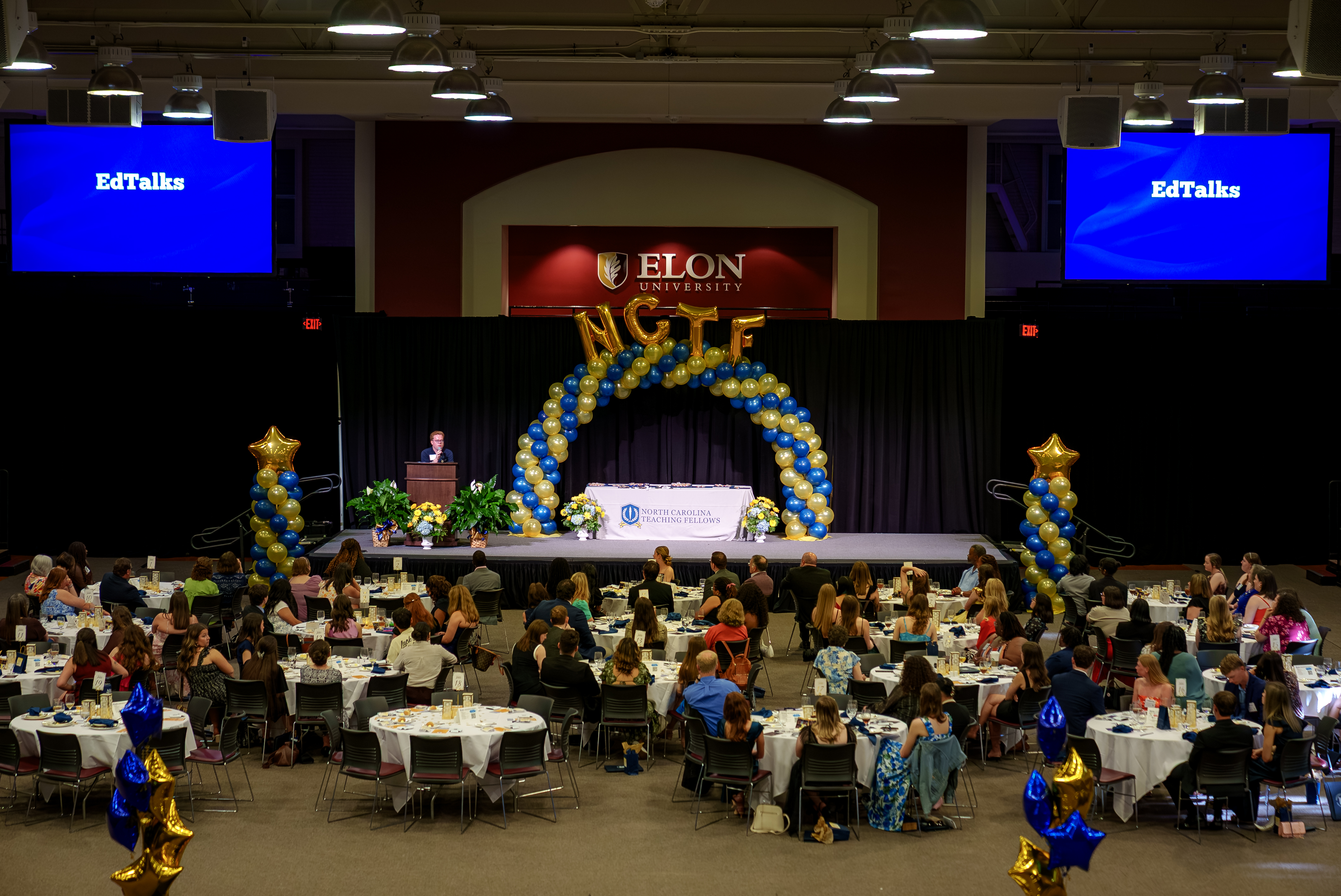  Wide view of a banquet hall at Elon University with guests seated at round tables facing a stage decorated with balloons and “EdTalks” screens.