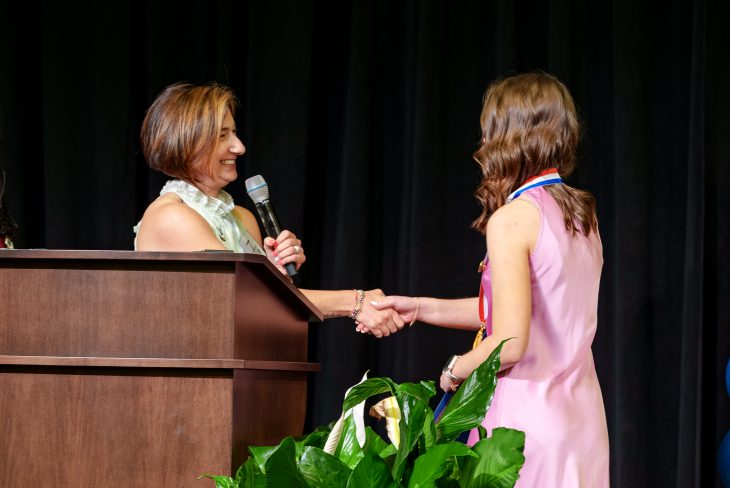 Two women shake hands on a stage. One stands behind a podium holding a microphone