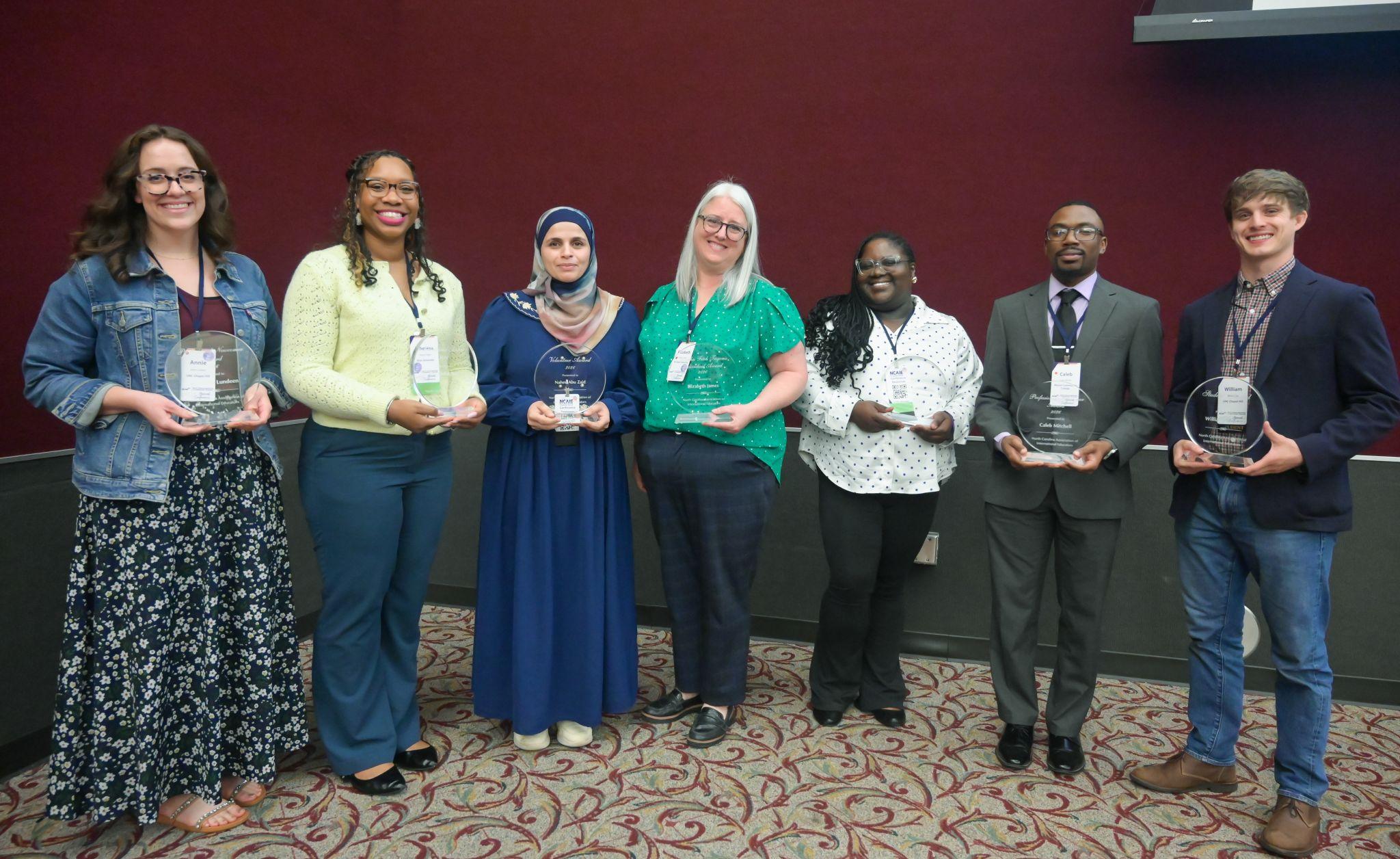 A group of seven people pose for a photo, all holding awards in hand