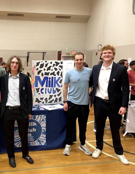 Three students stand in a gymnasium in front of a table display for “Milk Club,” featuring a cow-print sign and informational materials.