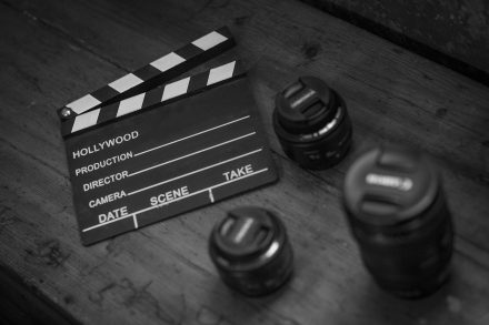 A film clapperboard labeled “Hollywood production” rests on a wooden surface beside several camera lenses.