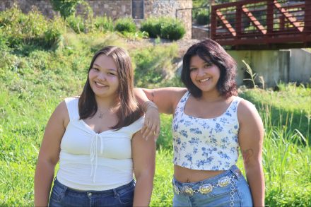 Two students, Reagan Wible (left) and Bella Pelini (right), smile while standing together outdoors in a sunny, natural setting. Bella rests her arm casually on Reagan’s shoulder, capturing a relaxed, friendly moment against a backdrop of greenery and a small bridge.