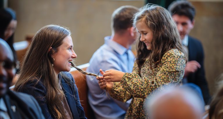 A young girl plays with her mother's hair before a ceremony in a courtroom. People are seated around them.