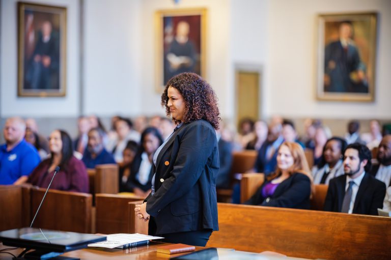 A woman smiles as she stands before a crowded courtroom.