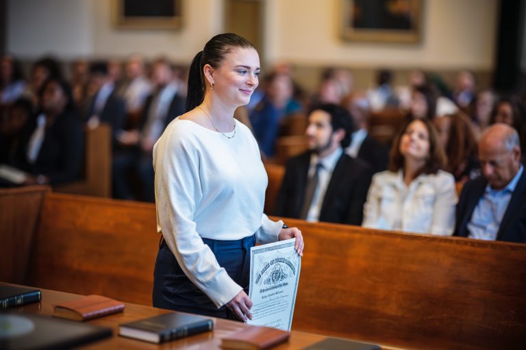 A woman walks through a crowded courtroom holding her law license, smiling.