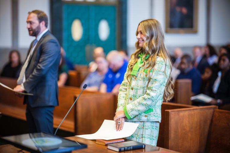 A woman in a floral suit smiles as a man speaks in a crowded courtroom