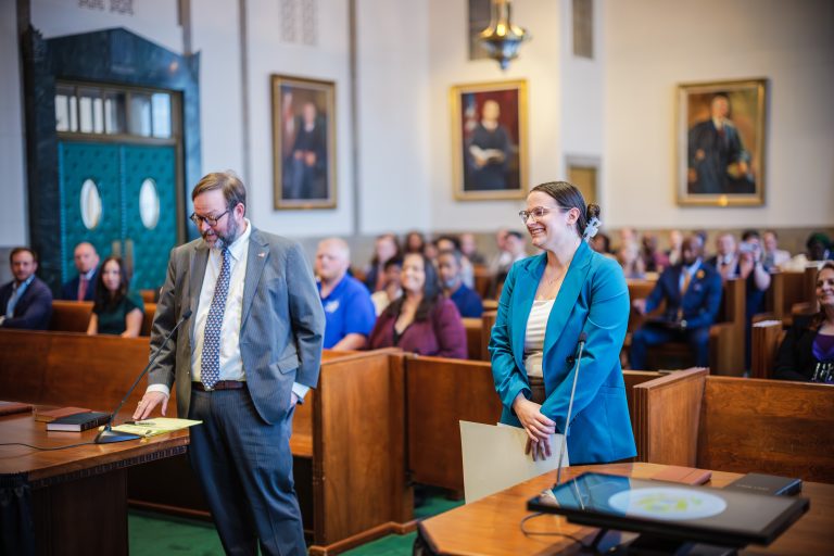 A woman in a blue suit smiles broadly as a man speaks to a crowded courtroom.