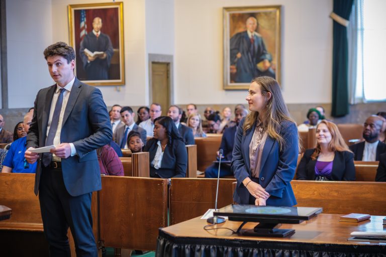 A man and woman stand across from each other in a crowded courtroom. He is looking directly ahead, speaking, she is smiling at him.