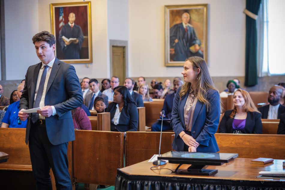 A man and woman stand across from each other in a crowded courtroom. He is looking directly ahead, speaking, she is smiling at him.