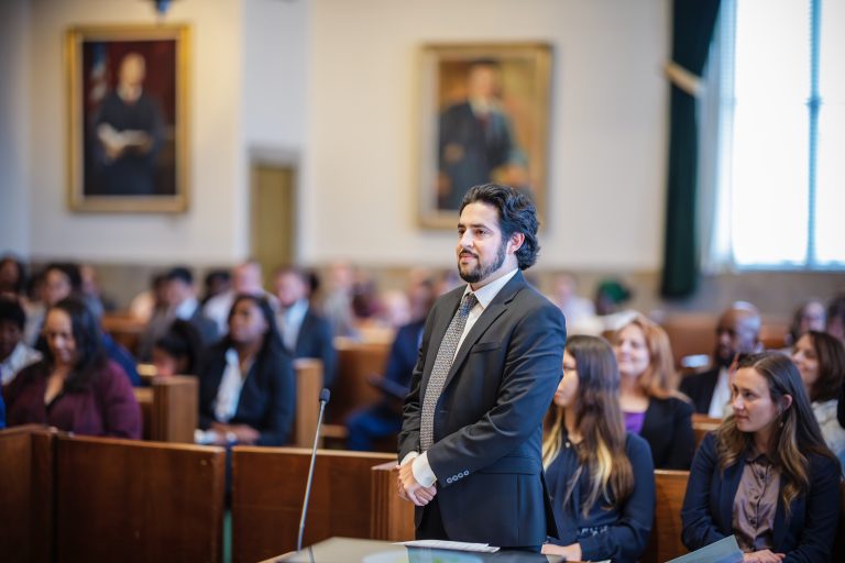 A man in a suit smiles confidently standing in front of a crowded courtroom.