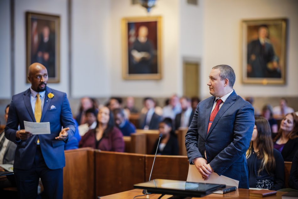 Two men stand in a crowded courtroom. The man on the left is speaking and looking at the man on the right, who is smiling.