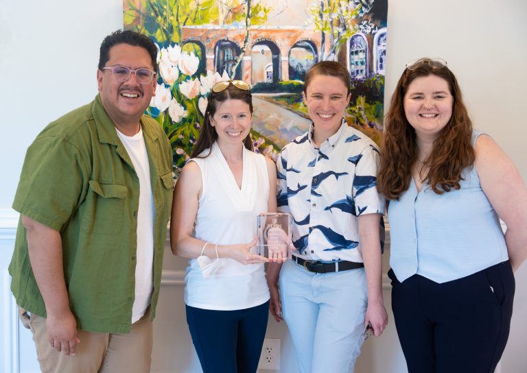Four individuals stand smiling indoors in front of a colorful painting during a recognition event. Two people in the center hold a glass award while posing together for the photo.