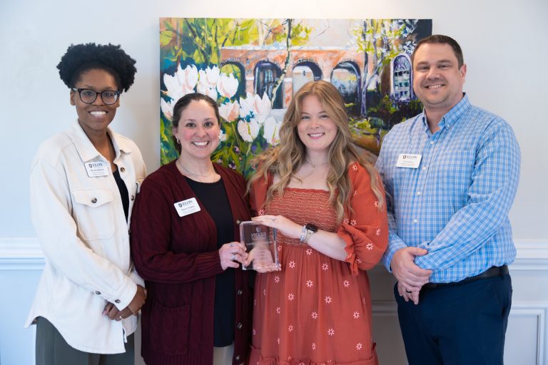 Four individuals stand smiling indoors in front of a colorful painting while posing with a glass award during a recognition event. Two people in the center hold the award as the group gathers for a commemorative photo.