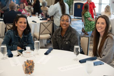 Three individuals sit at a round table smiling toward the camera during a recognition luncheon. Water glasses, folded napkins and a decorative jar sit on the table, while other attendees gather and converse in the background.