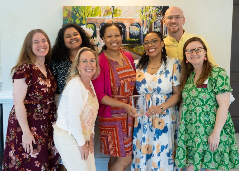 Seven individuals stand together smiling indoors in front of a colorful painting while posing with a glass award during a recognition event. Two people in the center hold the award as the group gathers for a celebratory photo.