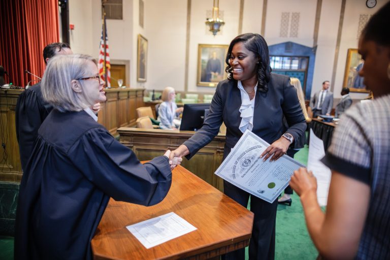 A judge wearing a robe shakes hands with a woman across a desk. They are smiling at each other.