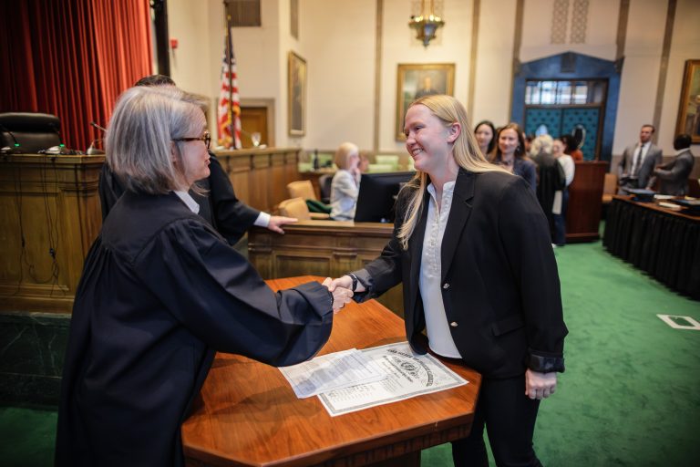 A judge in a robe shakes hands with a woman. A law license is beneath their hands on a desk in a courtroom.