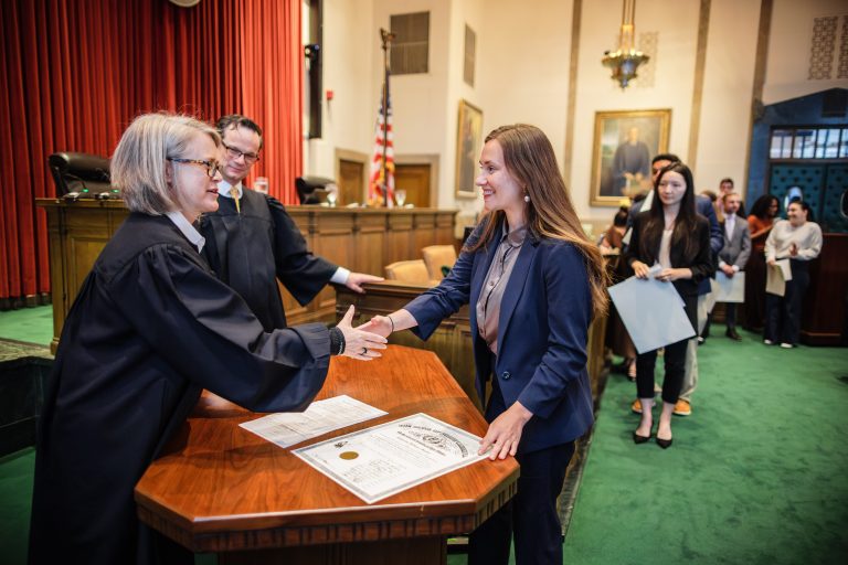 A judge wearing a robe shakes hands with another woman across a desk in a courtroom.