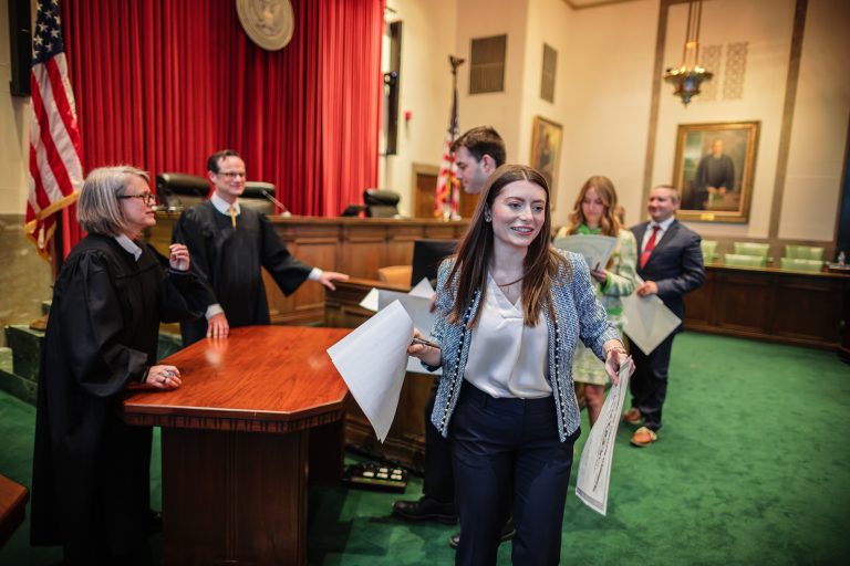 A woman smiling, holding paperwork. Judges and other law graduates are shown behind her.