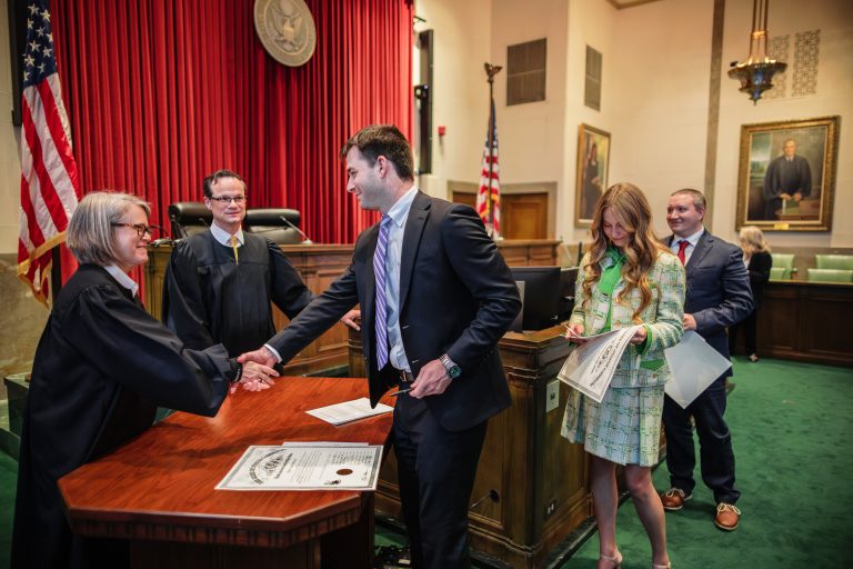 A judge wearing a robe shakes hands with a man over a desk. A law license is on the desk between them.