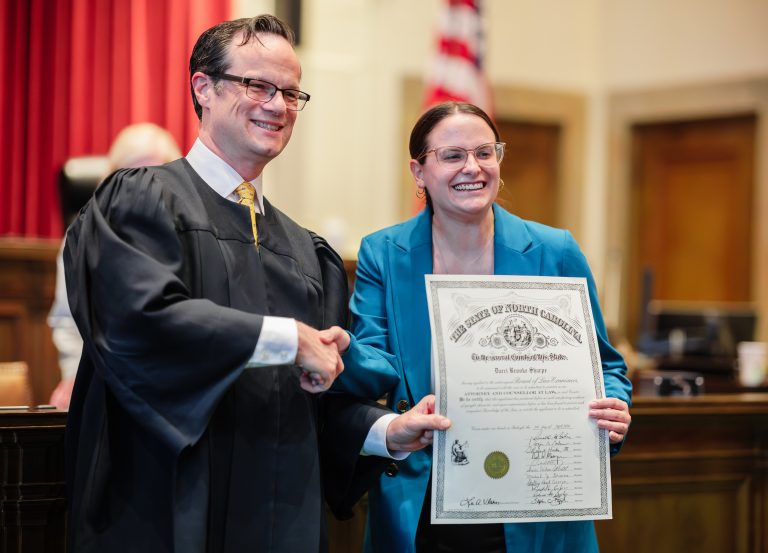 A judge and a woman pose for a photo. They are smiling and she is holding her law license.