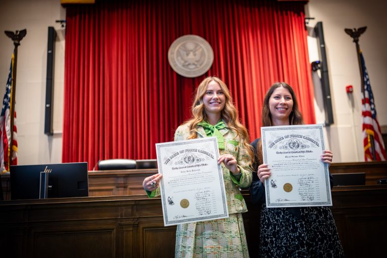 Two women stand with their law licenses, smiling. The federal seal is show behind them.