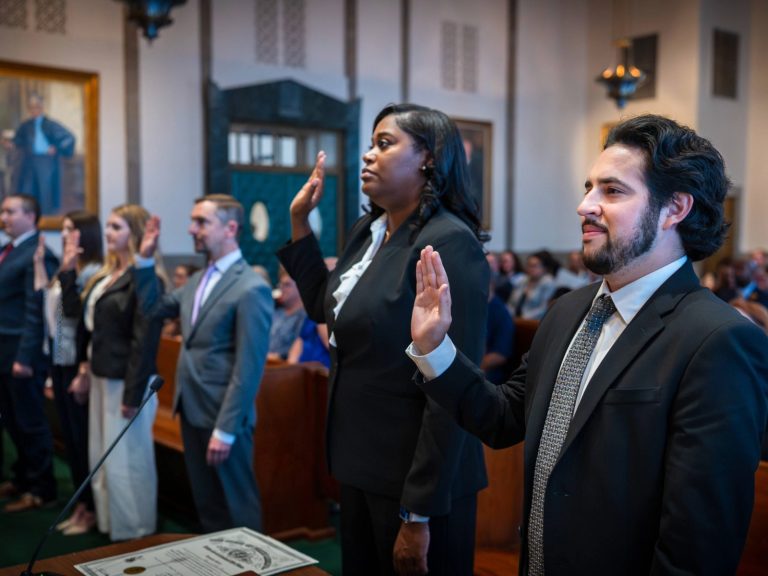 A group of people stand side by side with their right hands raised as they take an oath in a courtroom.