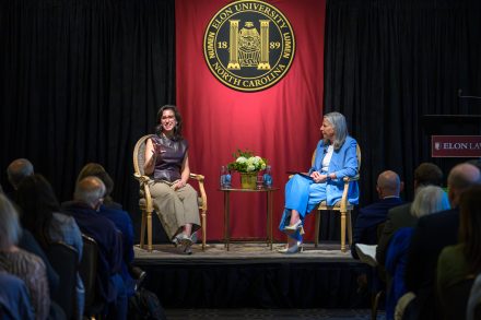 Two women seated on a stage with the ELon University seal behind them. The woman on the left is gesturing and smiling at the audience.