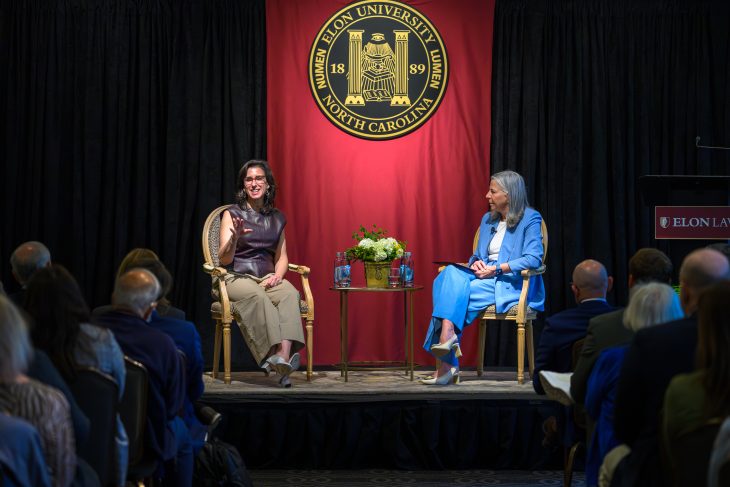 Two women seated on a stage with the ELon University seal behind them. The woman on the left is gesturing and smiling at the audience.