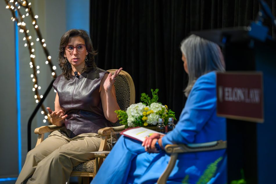 Two women seated on a stage. The woman on the left is gesturing and speaking. The woman on the right is listening.