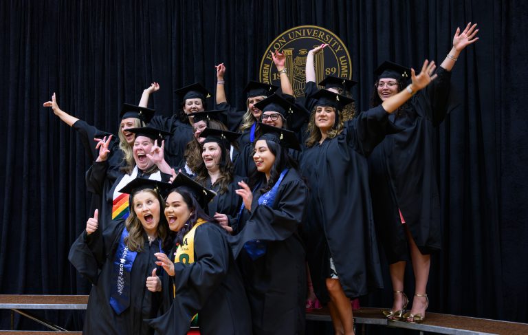A group of graduates in caps and gowns pose together with raised arms and smiles before Graduate Commencement.